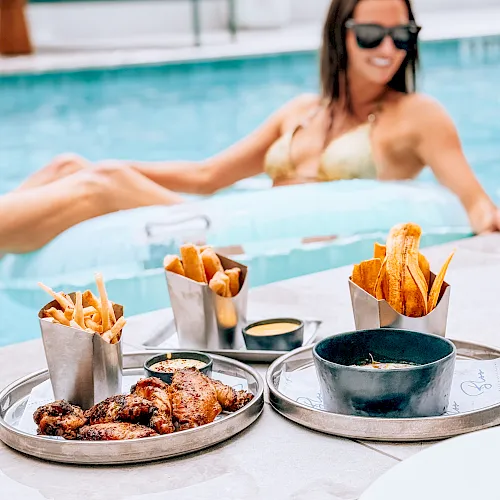 A woman in a pool wearing sunglasses, surrounded by a poolside food spread—fries, wings, sauces, and snacks.