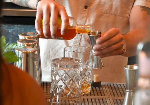 A bartender pours a drink with a smoky, focused vibe, using a shaker and glassware on a bar counter, in warm lighting.