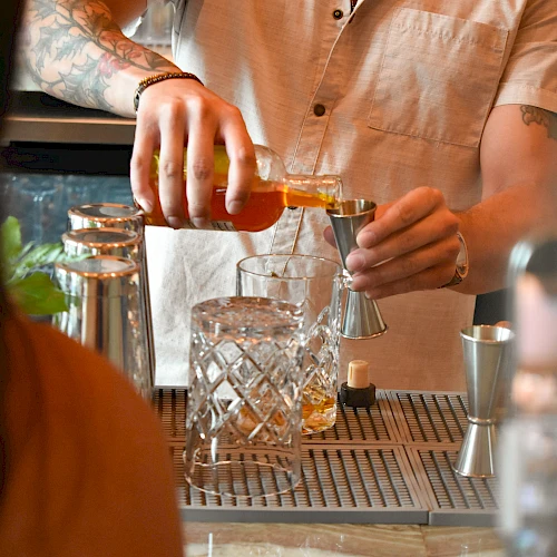A bartender pours a drink with a smoky, focused vibe, using a shaker and glassware on a bar counter, in warm lighting.