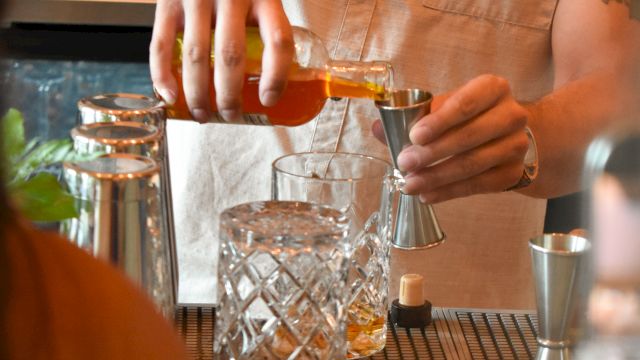 A bartender pours a drink with a smoky, focused vibe, using a shaker and glassware on a bar counter, in warm lighting.