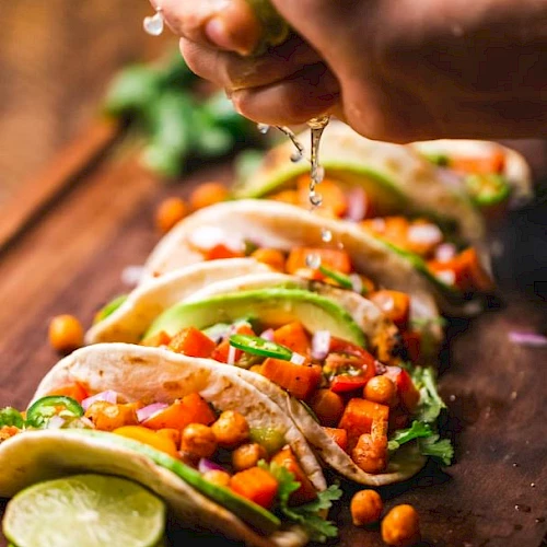 The image shows tacos filled with chickpeas, vegetables, and avocado, with a hand squeezing lime juice over them.