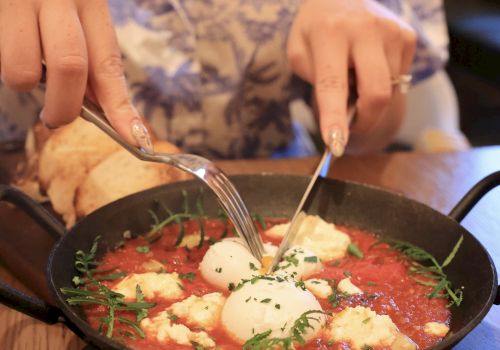 A person is eating shakshuka or soup in a skillet, using a spoon and fork, with a poached egg and herbs atop a tomato base.