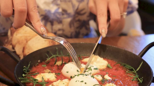 A person is eating shakshuka or soup in a skillet, using a spoon and fork, with a poached egg and herbs atop a tomato base.
