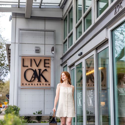 A stylish woman in a white dress stands outside a modern storefront with a wooden &ldquo;LIVE OAK&rdquo; sign, bright windows, and urban architecture.