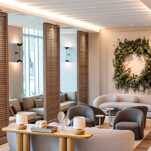 A modern, airy lounge with curved couches, wooden accents, soft lighting, a wreath wall decor, and cozy seating near tall windows