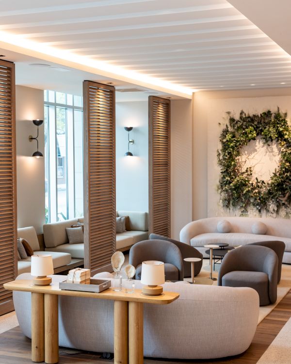 A modern, airy lounge with curved couches, wooden accents, soft lighting, a wreath wall decor, and cozy seating near tall windows