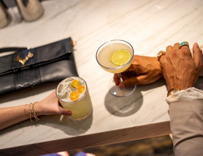 Two people clink cocktails at a counter: a pale drink with ice and lemon on the left, and a martini with a lemon twist on the right.