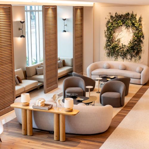 A modern, airy lounge with curved sofas, wooden dividers, soft seating, planters, a wreath wall art, and warm ambient lighting.
