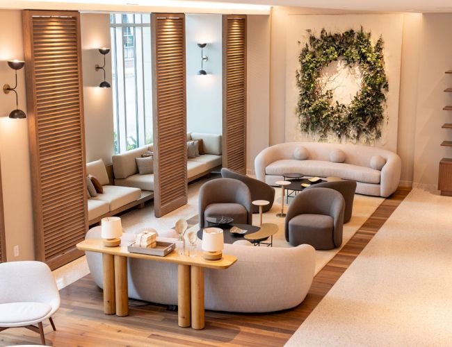 A modern, airy lounge with curved sofas, wooden dividers, soft seating, planters, a wreath wall art, and warm ambient lighting.