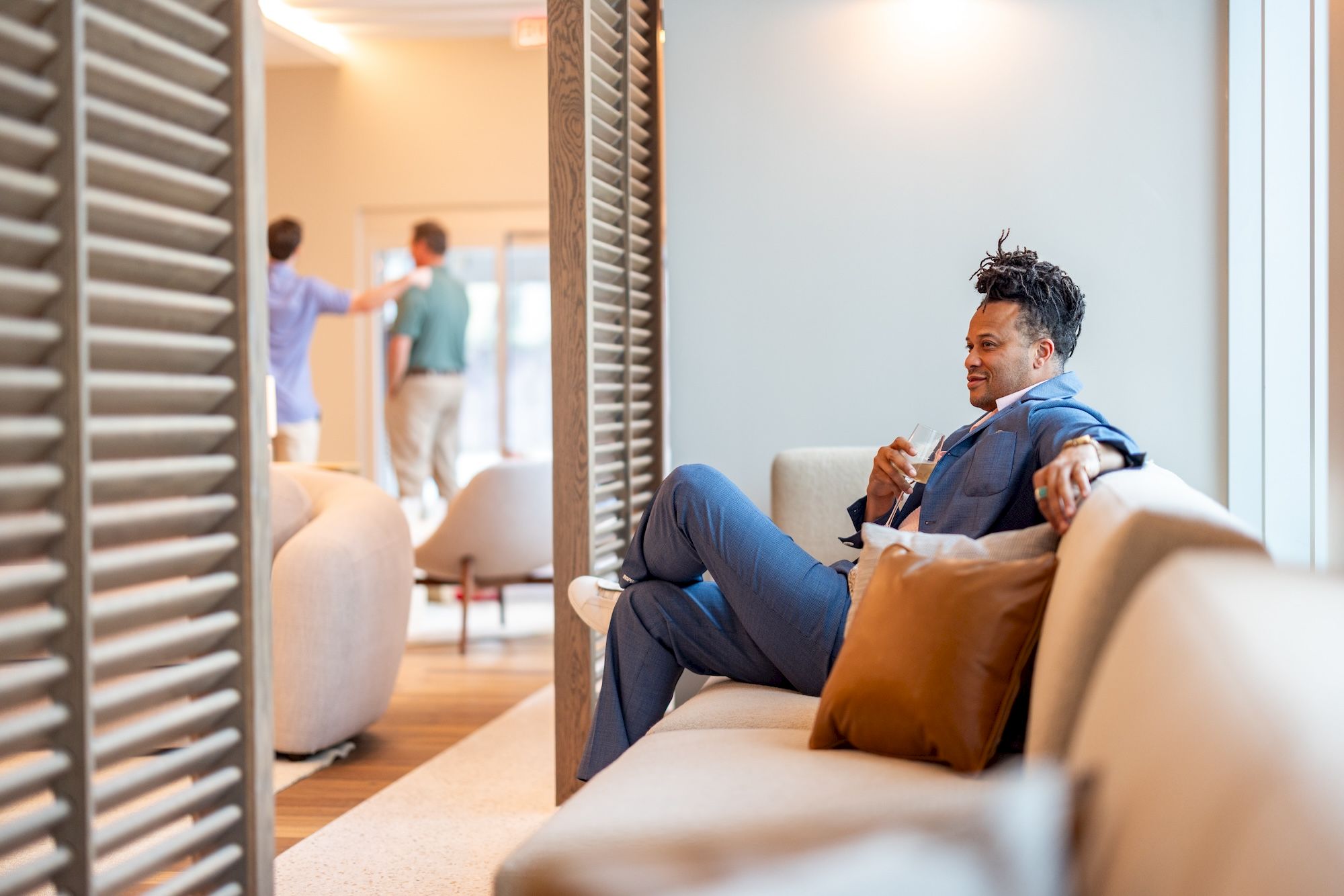 A man in a blue suit lounges on a beige couch with an orange pillow, relaxed, while colleagues chat in a bright, modern office in the background.