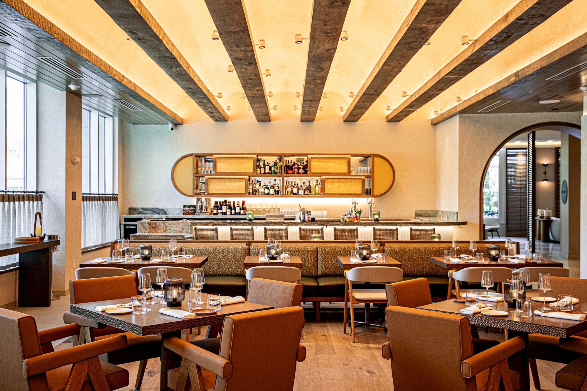 A stylish restaurant with wooden beams, warm lighting, and neatly set tables; a long shelf behind a bar area with glassware and bottles, inviting and cozy.