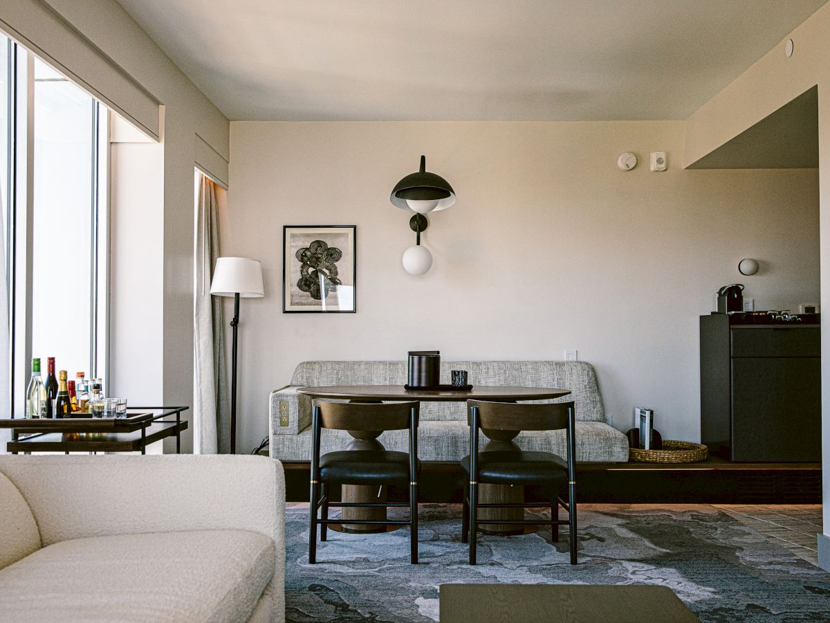 A modern living room with a small dining table and two chairs, a sofa on the left, a floor lamp, wall art, and a cabinet in the back, near a glass door.
