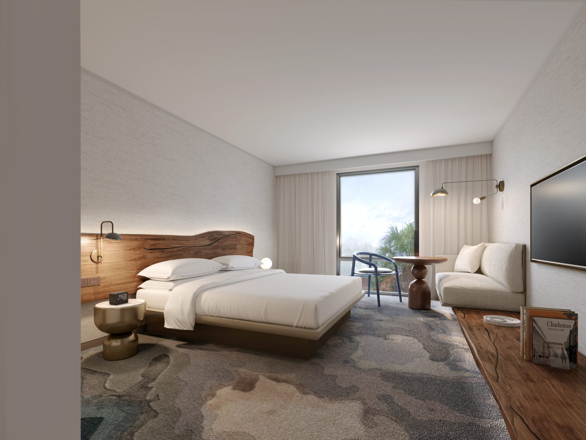A modern bedroom with a bed, nightstand, seating area, TV, and a view through sliding doors, featuring neutral tones and wood accents.
