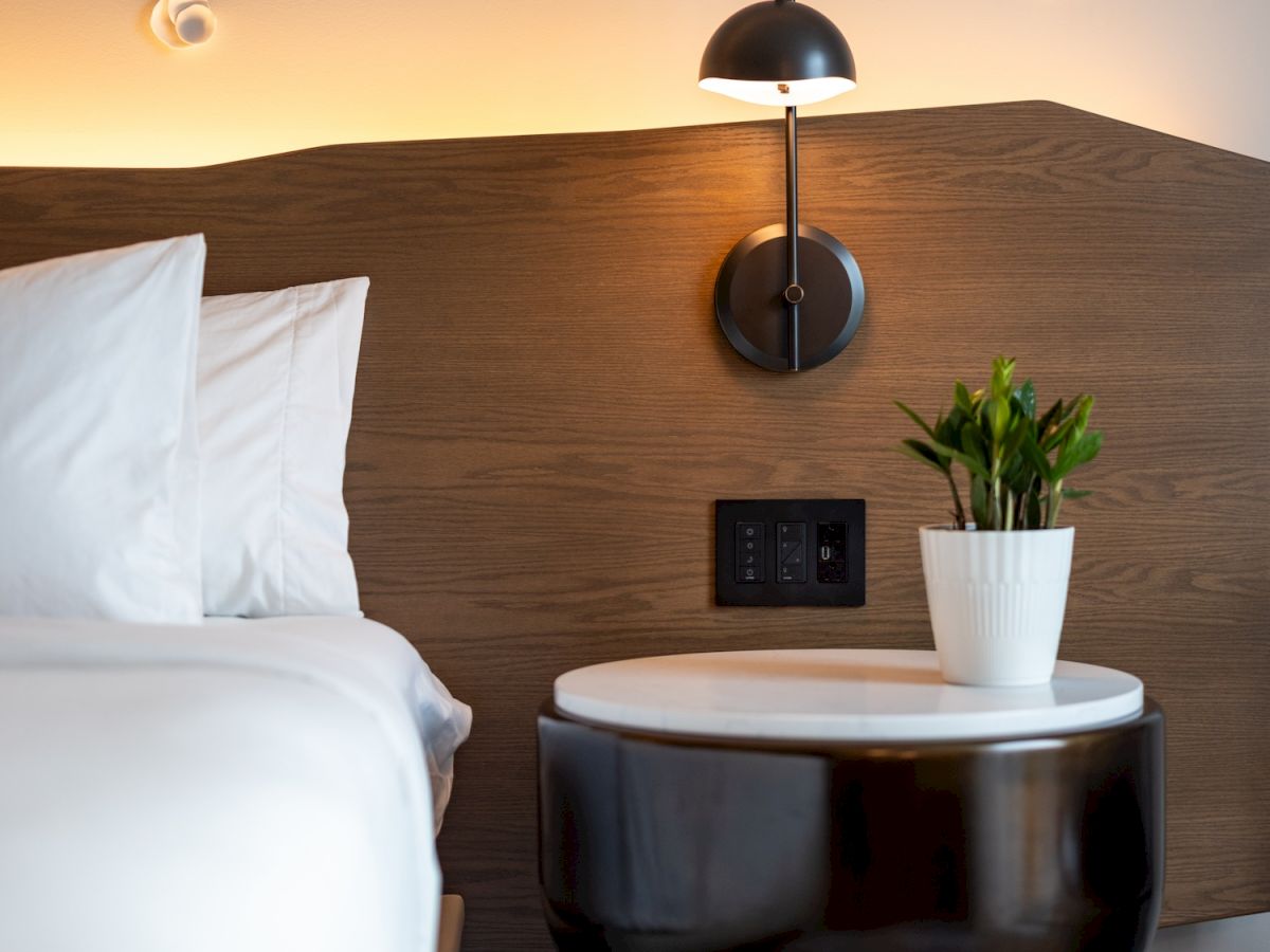 A stylish hotel room corner with a dark glossy nightstand, white marble top, potted plant, wall lamp, and soft reading light next to a white bed.