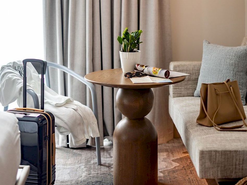 A hotel room scene with a round wooden table beside a sofa, a plant, a water bottle and snacks, a suitcase, and a coat/robe on the bed, cozy and modern.