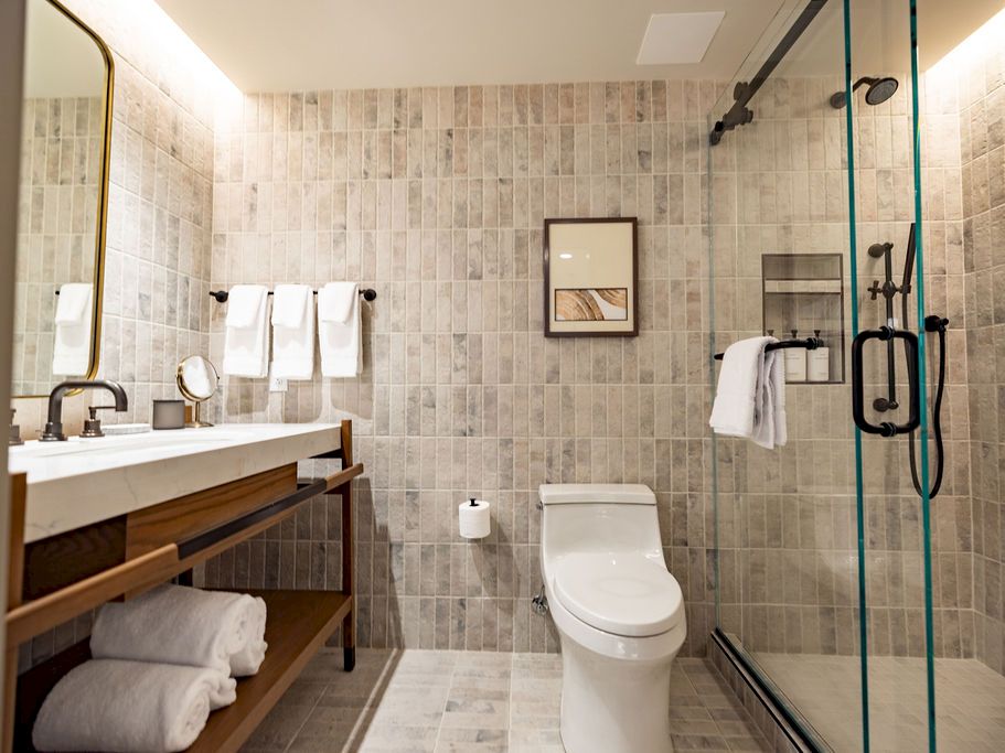 A modern bathroom with a double-sink vanity, wooden shelf, a wall mirror, towel racks, a toilet, and a glass-enclosed shower.