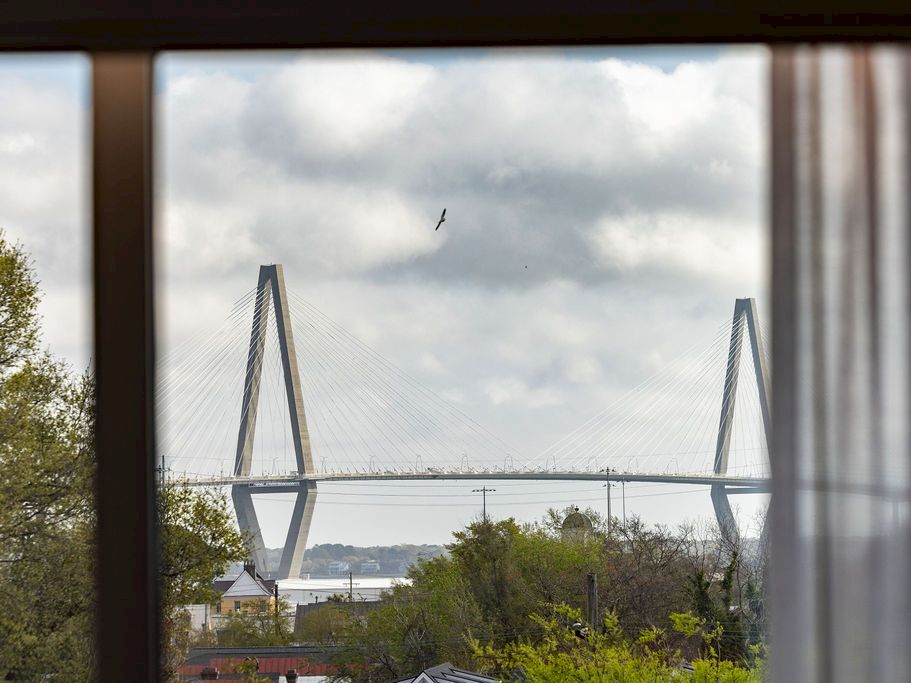 A distant suspension bridge framed by trees and a cloudy sky, with a lone bird soaring above.