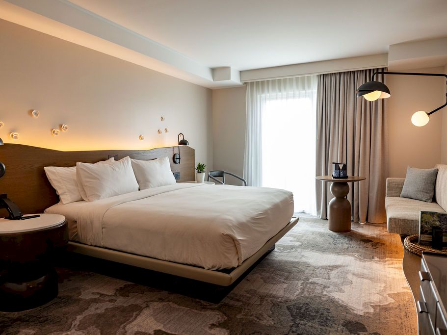 A modern hotel room with a large bed, soft lighting, a seating area, desk, and a balcony window, all in warm neutral tones.