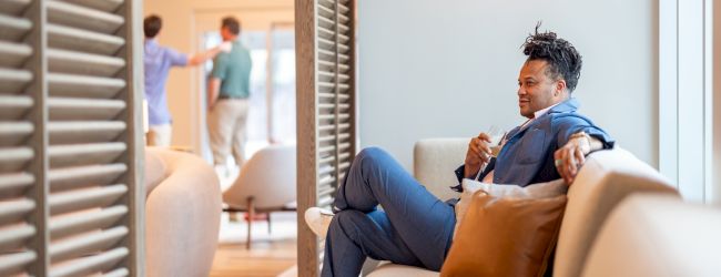 A cozy living room with a man lounging on a sofa, looking toward the doorway, while a child sits on the floor by a bookshelf.