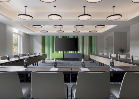 A modern conference room with long tables, overhead circular pendant lights, a large TV screen, and green-and-gray curtains.
