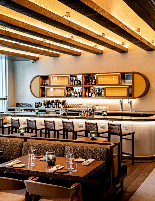 A stylish bar area with wooden beams, a long counter, and seating; warm lighting, shelves with bottles, and a modern dining setup. End.