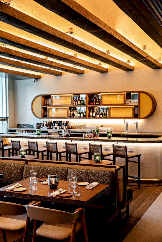 A stylish, modern restaurant interior with warm wood beams, a long bar, shelf/display behind the bar, and cozy dining tables with chairs. End sentence.