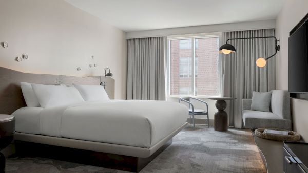 A modern hotel room with a large bed, grey and white decor, a seating area by the window, and contemporary lighting.