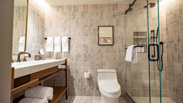A modern bathroom with a double vanity, open shelf towels, wall tiles, a wall-mounted toilet, and a glass-enclosed shower.