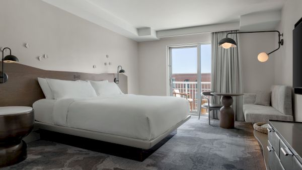 A modern hotel room with a large white bed, round side tables, a seating area, and a balcony view, bright and airy with neutral tones.