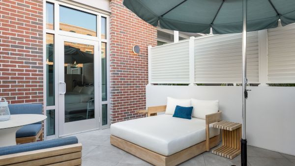 A modern outdoor patio lounge with a white daybed, blue pillows, a green umbrella, brick walls, and sliding glass doors, ready to relax.