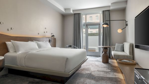A modern bedroom with a large white bed, gray curtains, bedside lamps, a lounge chair, and a flat-screen TV.