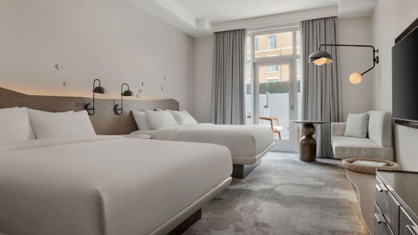 A modern hotel room with twin beds, gray curtains, a wall sconce, a sofa, and a large window overlooking the outside.
