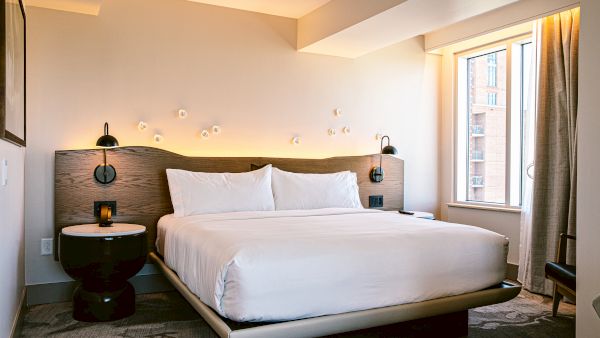 A modern hotel room with a large bed, white linens, two round black nightstands, wall lamps, and a window with curtains, warm lighting.