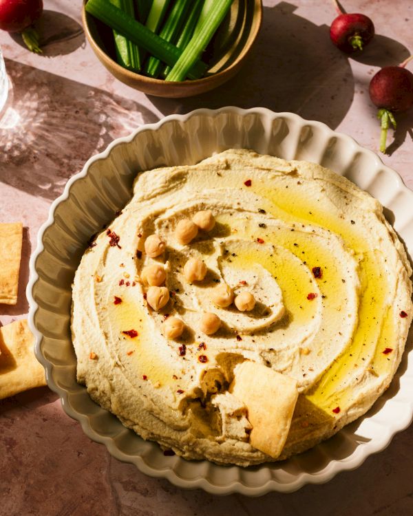 A bowl of hummus with olive oil and chickpeas, pita chips, green onions, radishes, and two drinks on a table.