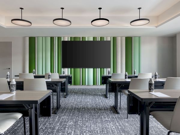 A modern meeting room with tables, chairs, hanging ceiling lights, green striped curtains, and a large wall display screen.