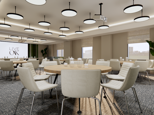 The image shows a modern conference room with round tables, neutral chairs, overhead lighting, and a screen displaying "LIVE ON."