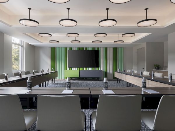 A modern conference room with long tables, chairs, hanging ceiling lights, a large TV, green curtains, and bottled water on desks.