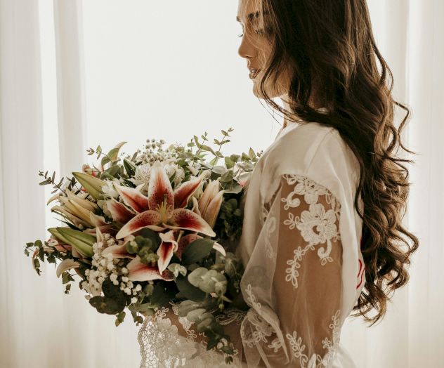 A woman in a bridal dress holds a bouquet of flowers in front of a softly lit curtain, creating a serene and elegant atmosphere.