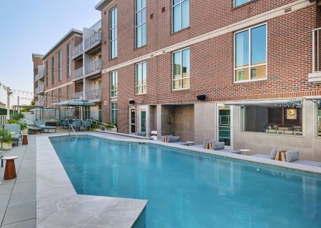 Modern brick apartment courtyard with a blue swimming pool, lounge chairs, string lights, and balconies.