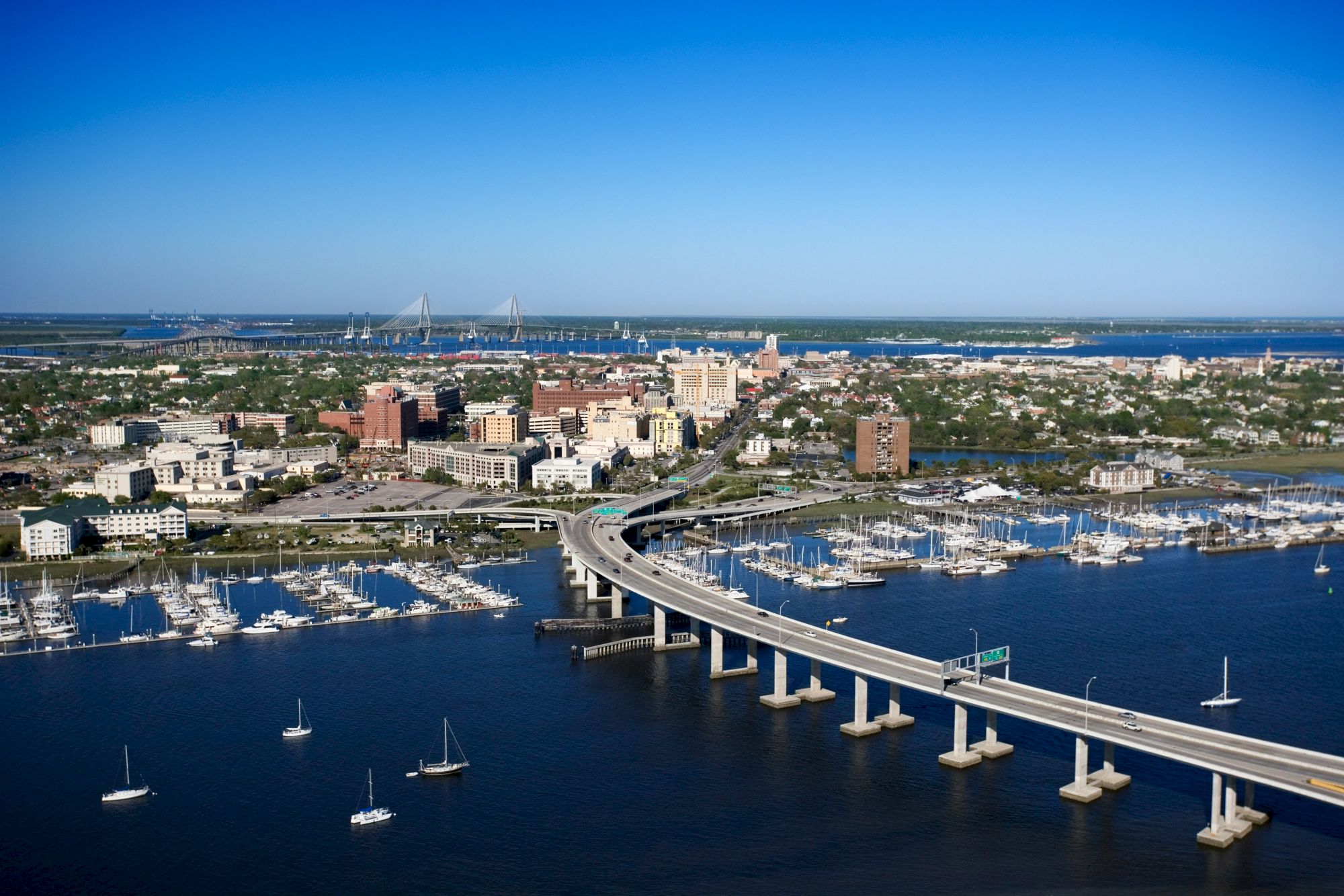 Aerial view of a coastal city with a bridge, marina, and buildings under a clear blue sky, showing a bustling waterfront scene.