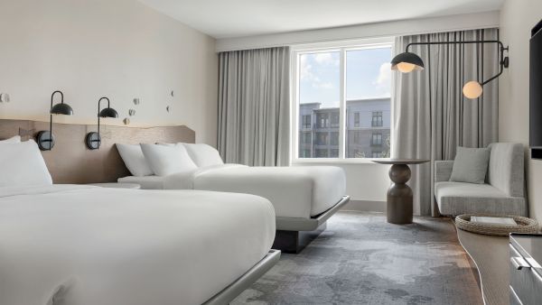 A modern hotel room with two queen beds, bedside lamps, a seating area, large window with curtains, and contemporary decor.