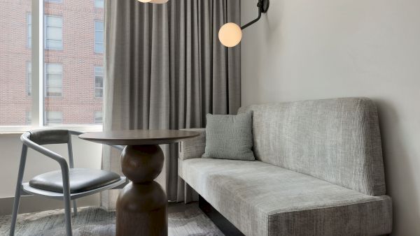 A modern hotel-like sitting area with a gray sofa, round wooden table, a gray chair, floor-to-ceiling curtains, and a stylish wall lamp by a large window.