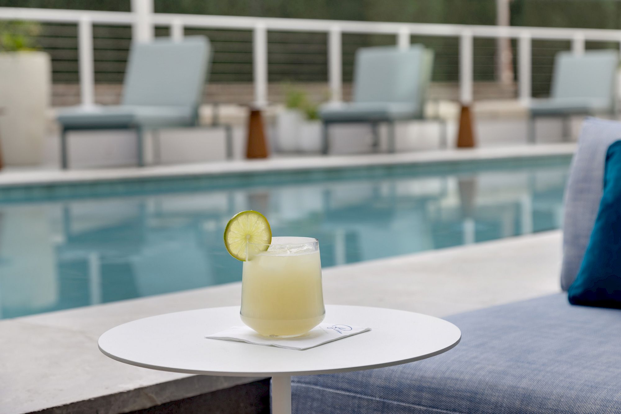 A refreshing pale yellow cocktail with a lime wheel on the rim sits on a round white plate beside a poolside lounge area.