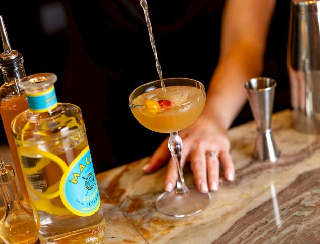 A bartender pours a cocktail into a coupe glass with a twist of lemon, next to bottles and bar tools on a marble counter.