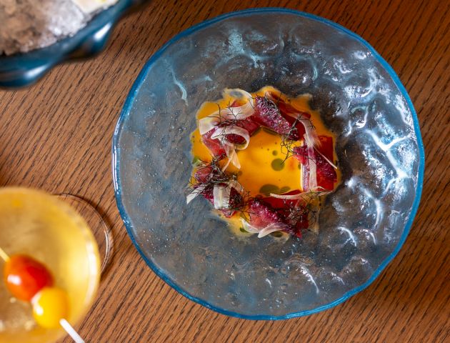 A close-up of a blue glass bowl with a colorful, messy egg yolk and vegetables, surrounded by other snacks on a wooden table, all viewed from above.