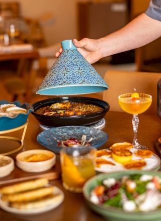 A table spread of tapas and small plates, grilled dishes, dips, cocktails, salad, and shared bowls; person lifting a blue ceramic dome to reveal food.