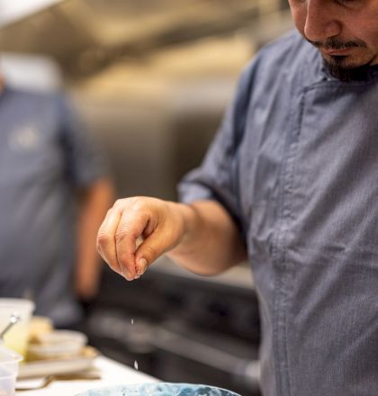 A chef sprinkles seasoning over a blue bowl with food, focused in a kitchen near a prep counter and blurred background.