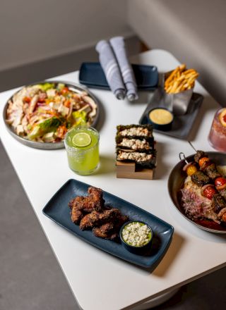 A spread of tapas and small plates: colorful veggies, dips, a bowl of greens, and grilled meats on stylish white plates.