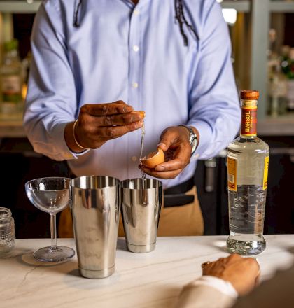 The image shows a bartender or chef pouring a drink into a glass, with a shaker and bar tools on the counter. Top it at 140 characters, always ending the sentence.