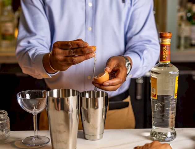 A bartender in a blue shirt pours a drink with metal shakers and a bottle of liquor on the bar, while a customer&rsquo;s hand reaches for ice.